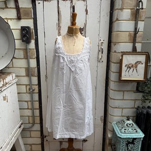 Può includere: Una camicia da notte vintage in cotone bianco con finiture in pizzo, esposta su un manichino di legno. La camicia da notte ha uno scollo tondo e una silhouette fluida. Lo sfondo comprende una porta bianca, un muro di mattoni e elementi decorativi antichi.