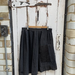 May include: A dark gray skirt with a striped pattern and a patch pocket. The skirt is hanging on a wooden hanger against a white wooden door.