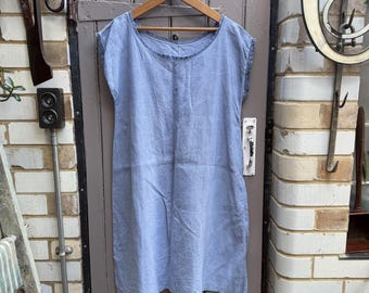 May include: A light blue, short-sleeved dress hangs on a wooden hanger against a weathered gray door. The dress has a simple, relaxed design, with a round neckline and a slightly textured appearance. The overall aesthetic is casual and comfortable.