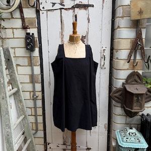Könnte beinhalten: Ein schwarzes Kleid mit quadratischem Ausschnitt, das auf einer Holzpuppe präsentiert wird. Das Kleid hat ein schlichtes, ärmelloses Design. Die Puppe steht vor einer weißen Tür mit der Nummer 21, mit einer Backsteinmauer im Hintergrund.