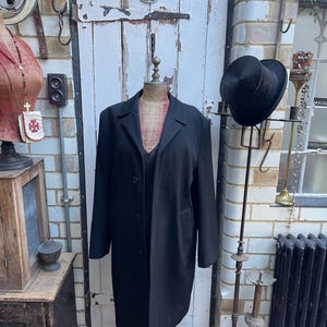 May include: A black long-sleeved coat with a single button closure, displayed on a mannequin against a white brick wall.