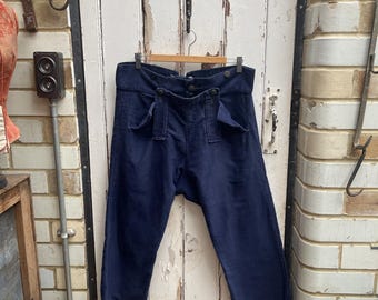 Puede incluir: Un par de pantalones de pierna ancha de color azul oscuro con cierre de botón y varios bolsillos. Los pantalones están colgados de una percha de madera contra una puerta de madera blanca.