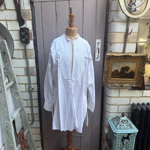 May include: A long-sleeved white shirt featuring a mandarin collar, a central front placket, and side slits at the hem. The shirt is displayed on a wooden mannequin torso, showcasing a loose, straight-cut fit with simple buttoned cuffs.