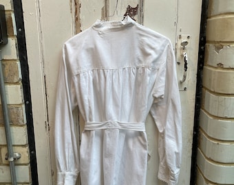 May include: White cotton dress with a ruffled collar and long sleeves. The dress has a tie belt at the waist and a white trim around the cuffs.