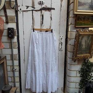 May include: A white embroidered skirt hangs on a wooden hanger against a distressed white door. The skirt has detailed eyelet and floral patterns, with a tiered, ruffled hem. The door has a metal number 21 plate.