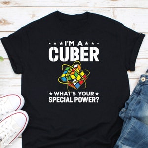May include: Black t-shirt with white text that reads "I'm a Cuber" and "What's your special power?"  A colorful Rubik's Cube is in the center of the shirt.