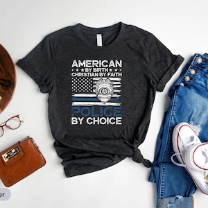 May include: Dark heather grey t-shirt with a distressed American flag design and the text "American by Birth, Christian by Faith, Police by Choice".