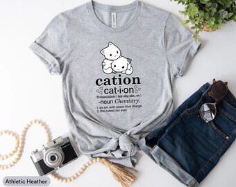 May include: Gray t-shirt with a cute cartoon illustration of two white kittens hugging. The shirt has the word "cation" written in black text with the definition "cat-i-on" and a pronunciation guide. The definition continues with "noun Chemistry. 1. an ion with paws-itive charge 2. the cutest ion ever"