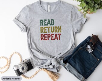 Puede incluir: Una camiseta gris con el texto "READ RETURN REPEAT" en una fuente de estilo vintage. El texto está en verde, amarillo y rojo.