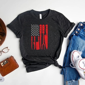 May include: Dark heather grey t-shirt with a red, white, and blue American flag design featuring a set of kitchen knives. The shirt is displayed on a white surface with a pair of jeans, a camera, and a small potted plant.