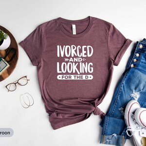 May include: A maroon t-shirt with the text "IVORCED AND LOOKING FOR THE D" in white. The shirt is laid flat on a white surface with a pair of jeans and white trainers.