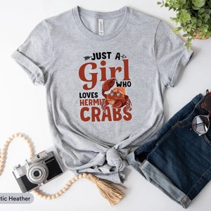 May include: Athletic heather gray t-shirt with the text "Just a Girl Who Loves Hermit Crabs" in brown and white. The shirt features a cartoon hermit crab graphic. The shirt is paired with blue denim shorts and sunglasses.