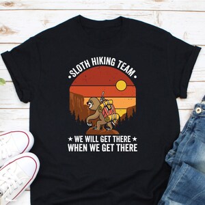 May include: Black t-shirt featuring a retro-style graphic of a sloth hiking with a backpack. The text reads "Sloth Hiking Team. We will get there when we get there."