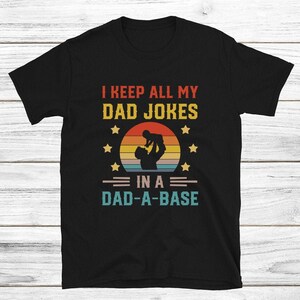 May include: Black t-shirt with a retro-style sunset graphic and the text "I keep all my dad jokes in a dad-a-base".