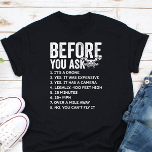 May include: Black t-shirt with white text that reads "BEFORE YOU ASK" and a drone graphic. The text lists eight facts about drones, including that they are expensive, have cameras, and can fly over a mile away.
