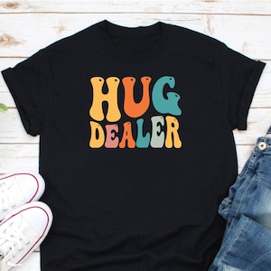 May include: Black t-shirt with the text "HUG DEALER" in colorful retro-style lettering.