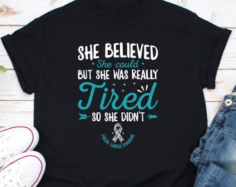 May include: Black t-shirt with a teal blue graphic that reads "She Believed. She could. But she was really Tired. So she didn't." with a teal blue ribbon and the words "Ehlers-Danlos Syndrome" below.