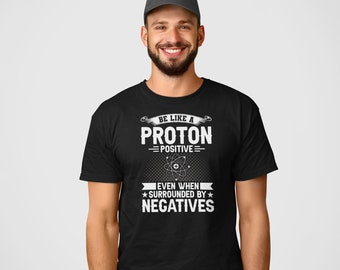 May include: Black t-shirt with a white graphic that says "Be like a proton - positive - even when surrounded by negatives". The graphic also includes a stylized atom.