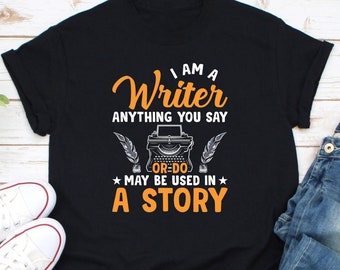 May include: Black t-shirt with a white and orange graphic design. The graphic features a vintage typewriter with the text "I am a Writer Anything you say or do may be used in a story."