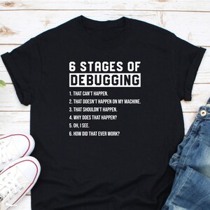 May include: Black t-shirt with white text that reads "6 Stages of Debugging" and lists the stages: "That can't happen", "That doesn't happen on my machine", "That shouldn't happen", "Why does that happen?", "Oh, I see", and "How did that ever work?"