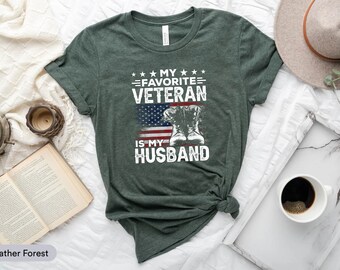 May include: A green t-shirt with the text "My Favorite Veteran is My Husband" and a graphic of an American flag and military boots.