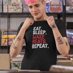 May include: A black t-shirt with the text "EAT. SLEEP. MAKE BEATS. REPEAT." in red and white lettering. The person wearing the shirt has pink hair and tattoos on their arms.