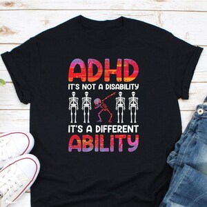 May include: Black t-shirt with a colorful graphic design that says "ADHD It's not a disability It's a different ability". The graphic features a skeleton doing the dab dance and four other skeletons standing in a line.