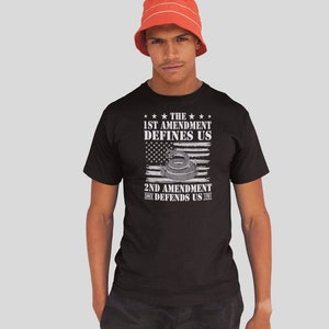 May include: A black t-shirt with a white graphic design featuring an American flag with a gun silhouette and the text "The 1st Amendment Defines Us. 2nd Amendment Since Defends Us 1791."
