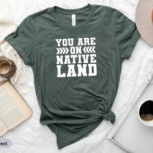 May include: A dark green t-shirt with the text "YOU ARE ON NATIVE LAND" in white lettering. The shirt is tied in a knot at the bottom.
