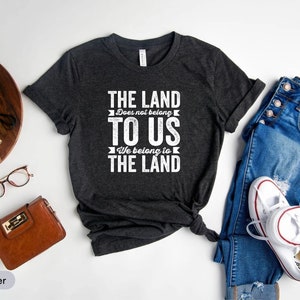 May include: Dark heather grey t-shirt with a white graphic print that reads "THE LAND Does not belong TO US We belong to THE LAND".