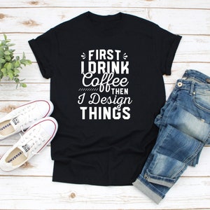 May include: Black t-shirt with white text that reads "First I drink coffee then I design things". The shirt is laid flat on a wooden surface with a pair of white sneakers and blue jeans.