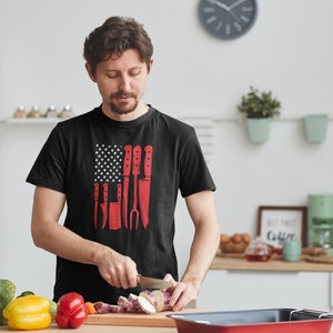 May include: A person wearing a black t-shirt with a red, white, and blue American flag design featuring a row of kitchen knives. The person is cutting a purple yam on a wooden cutting board in a kitchen.