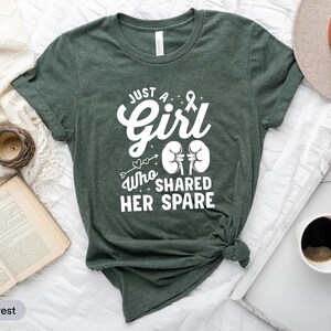 May include: A green t-shirt with white text that reads "Just a Girl Who Shared Her Spare". The text is in a stylised font and includes a heart, arrow, and a kidney symbol.