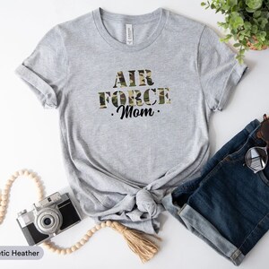 May include: A gray t-shirt with the text "AIR FORCE Mom." in black letters with a camouflage pattern. The shirt is tied in a knot at the bottom.