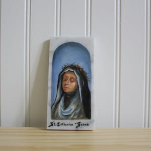 Può includere: Una placca di marmo bianco con un dipinto di Santa Caterina da Siena. Il dipinto la raffigura con una corona di spine e una veste blu e nera. Il testo "St. Catherine "Siena" è scritto sotto il dipinto.