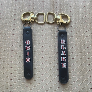 May include: Two black leather keychains with the names "ORIO" and "BLAKE" in red and white lettering. The keychains are attached to brass-colored clasps and rings. The background is a textured, patterned surface.