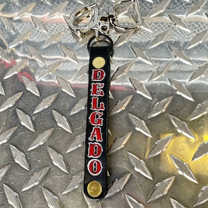 May include: Black leather keychain with a silver clip and the name "DELGADO" in red lettering.