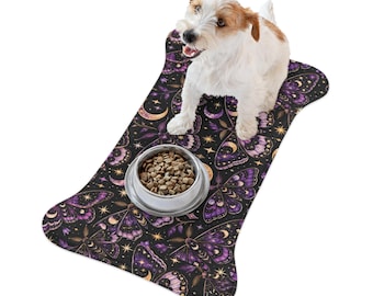 May include: A bone-shaped pet feeding mat featuring a dark purple and black pattern of moths, crescent moons, and stars. A silver metal bowl filled with dry brown kibble sits on the mat, with a small white and tan dog standing on top.