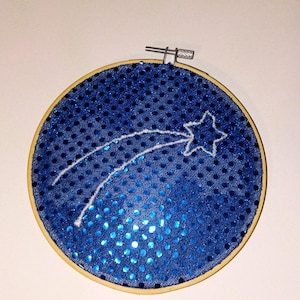 May include: A round embroidery hoop featuring a blue sequined fabric background. A white shooting star design is stitched across the fabric. The hoop is made of wood and has a metal clasp.
