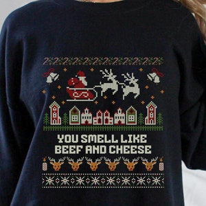 Op de afbeelding: Zwarte kersttrui met een wit, rood en groen pixelpatroon. De trui heeft een kerstman in een slee getrokken door rendieren, huizen en de tekst "You smell like beef and cheese".