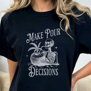 May include: Black t-shirt with a white graphic of a skeleton drinking from a coconut, with the text "Make Pour Decisions". The design includes palm leaves and stars. The shirt is a casual wear item.