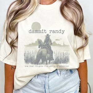 May include: Cream-colored t-shirt with a graphic of a cowboy on a horse, with the text "dammit randy" above and "now that I'm gone I've got your attention" below. The shirt is a relaxed fit.
