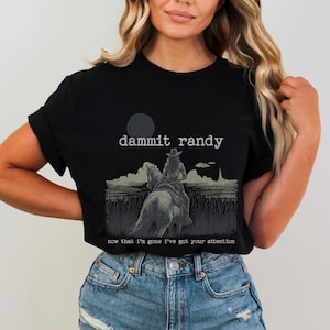 May include: Black t-shirt featuring a graphic of a person riding a horse, with the text "dammit randy" above and "now that I'm gone I've got your attention" below. The design is in shades of gray and white.
