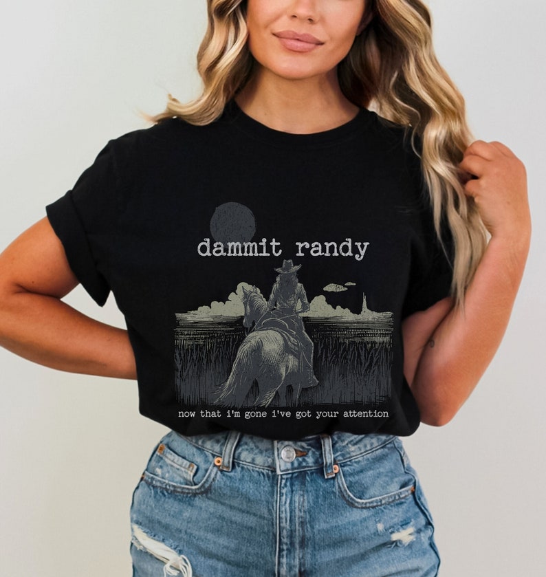 May include: Black t-shirt featuring a graphic of a person riding a horse, with the text "dammit randy" above and "now that I'm gone I've got your attention" below. The design is in shades of gray and white.