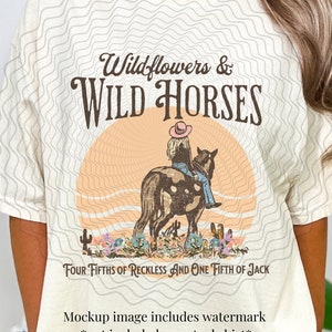 K&ouml;nnte beinhalten: Ein beiges T-Shirt mit einem Grafikdesign einer Frau, die auf einem Pferd vor einem Sonnenuntergang reitet. Der Text "Wildflowers & WILD HORSES" befindet sich &uuml;ber dem Bild und der Text "FOUR FIFTHS OF RECKLESS AND ONE FIFTH OF JACK" unter dem Bild.