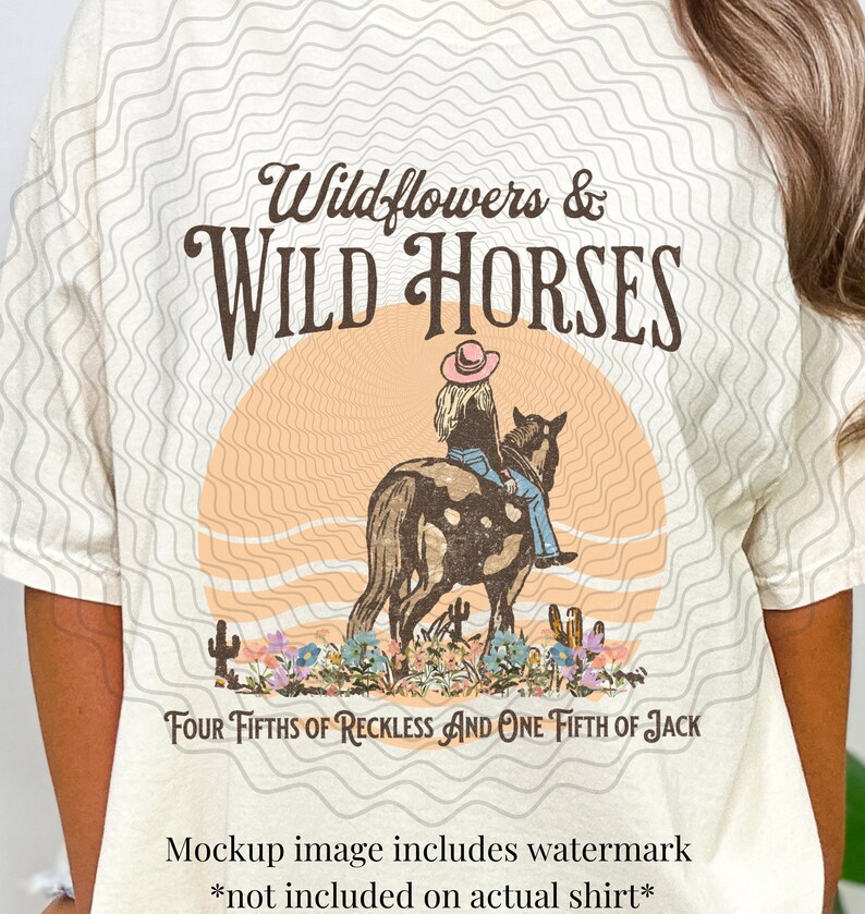 K&ouml;nnte beinhalten: Ein beiges T-Shirt mit einem Grafikdesign einer Frau, die auf einem Pferd vor einem Sonnenuntergang reitet. Der Text "Wildflowers & WILD HORSES" befindet sich &uuml;ber dem Bild und der Text "FOUR FIFTHS OF RECKLESS AND ONE FIFTH OF JACK" unter dem Bild.