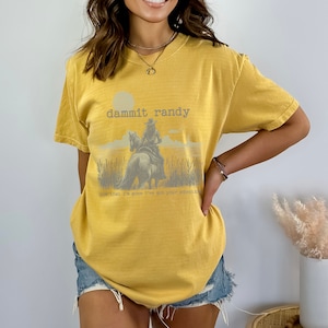 May include: Mustard yellow t-shirt with a graphic of a cowboy on a horse, with the text "dammit randy" and "now that I'm gone I've got your attention". The shirt is a short-sleeved, casual style.