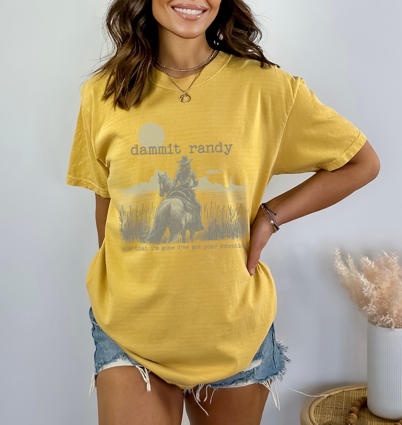 May include: Mustard yellow t-shirt with a graphic of a cowboy on a horse, with the text "dammit randy" and "now that I'm gone I've got your attention". The shirt is a short-sleeved, casual style.