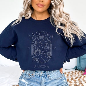 May include: Navy blue sweatshirt with a white graphic design of a desert landscape with mountains, cacti, and a road. The graphic is in a circle with the text "SEDONA" above the circle and "ARIZONA" below the circle. The numbers "19" and "02" are on either side of the circle.