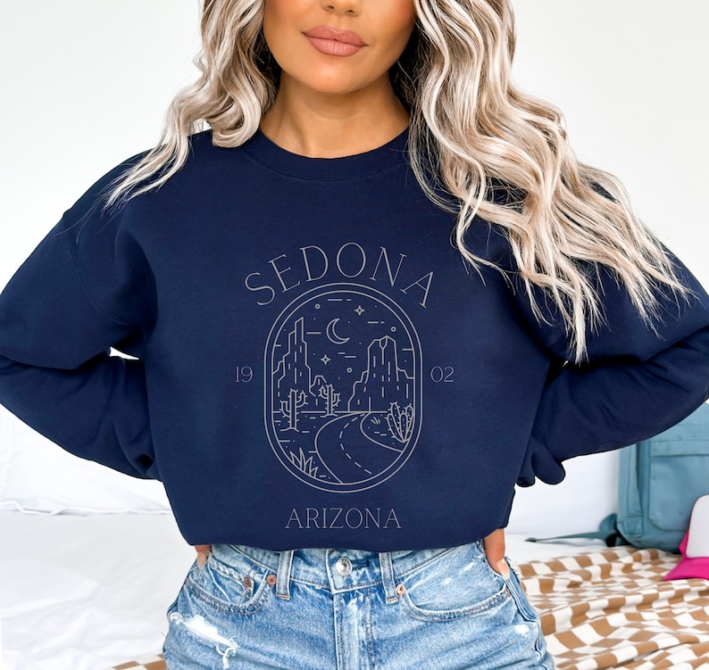 May include: Navy blue sweatshirt with a white graphic design of a desert landscape with mountains, cacti, and a road. The graphic is in a circle with the text "SEDONA" above the circle and "ARIZONA" below the circle. The numbers "19" and "02" are on either side of the circle.